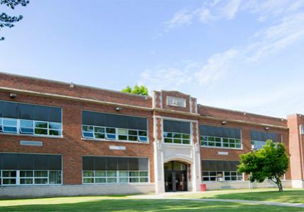 Exterior of two story 1920's high school with large trees and green lawn out front