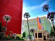 Outside view of a light green building with a US flag hanging and purple flowers framing the photo