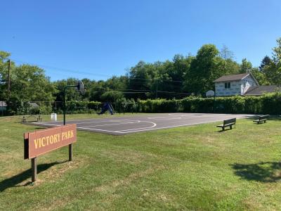 Brown and yellow Victory park sign on green grass, next to a basketball court and playground equipment