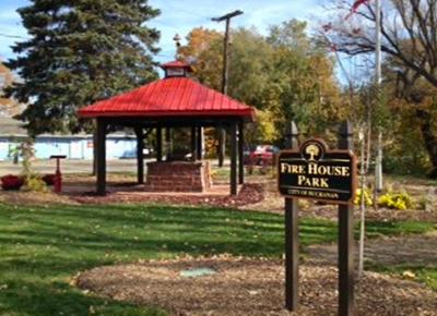 A red roofed structure housing historic firehouse bell and a sign that says firehouse park