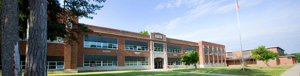 Exterior of two story 1920's high school with large trees and green lawn out front